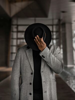 Elegant photo of a man in a formal coat and fedora hat posing indoors.